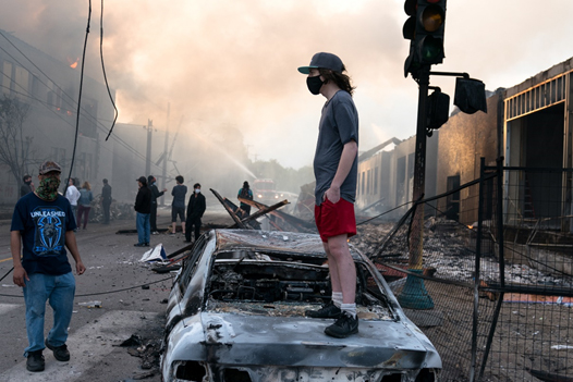 A young, masked man, standing on top of a burned-out car in a street riot.